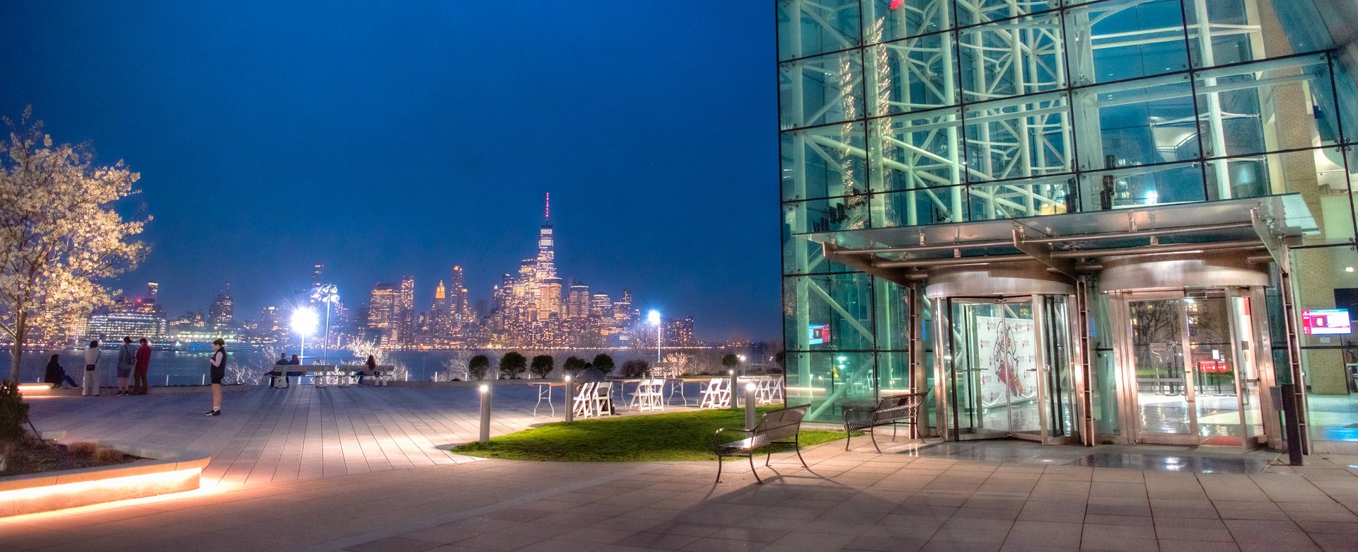 The glass entrance of the Babbio Center at night, with the lit Manhattan skyline across the Hudson River