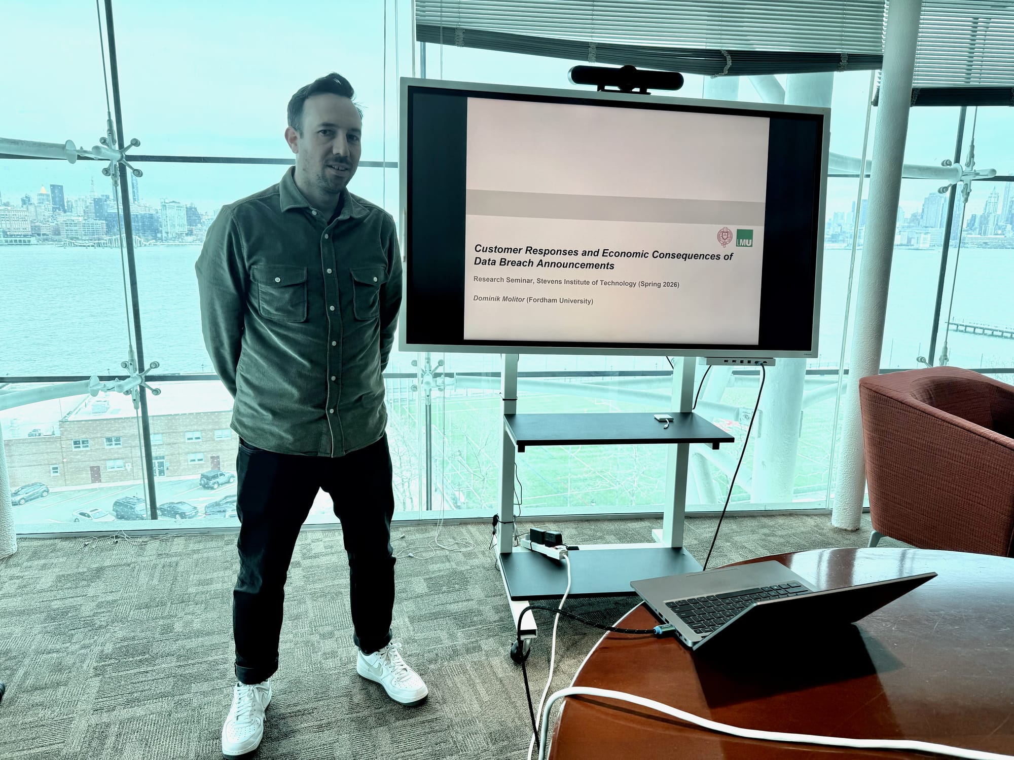 Dominik Molitor stands beside a screen showing the title slide of his talk, with the river and Manhattan behind the windows