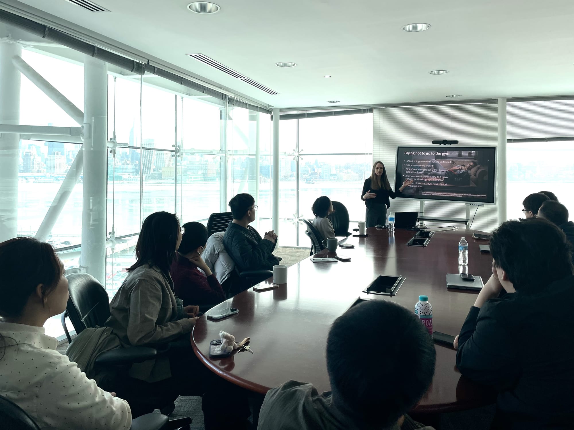Sagit Bar-Gill presents to an audience seated around a conference table, with the Manhattan skyline in the windows behind her